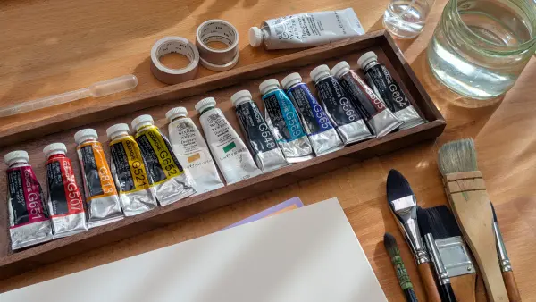 Gouache paint tubes, brushes and a water jar on a wooden table